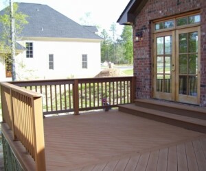 Wooden deck leading to a brick house with glass doors, railing on one side, and a view of neighboring house and tree in the background.