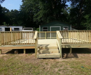 Wooden deck with railings and stairs in a grassy yard, fronting two small buildings surrounded by lush trees.