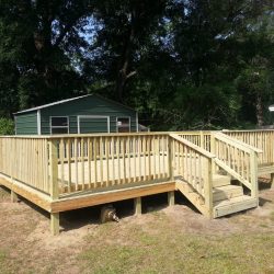 A newly constructed wooden deck with railings and stairs in a backyard, next to a green shed and surrounded by trees.