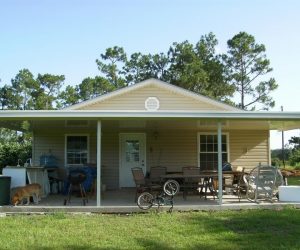 Single-story house with a wide porch, outdoor furniture, a bicycle, and a dog. Trees and green lawn surround the house.